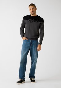 Grauer und schwarzer Langarm-Sweatshirt mit dem Aufdruck "GUESS EST.1981" auf der Vorderseite, kombiniert mit hellblauen, locker sitzenden Jeans und schwarzen Turnschuhen.