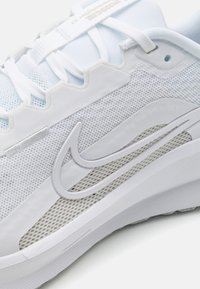 Încălțăminte sport albă, realizată din materiale din plasă și sintetice, cu o talpă texturată și un logo stilizat proeminent pe lateral.