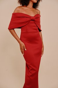 Žena oblečená v priliehavých, bezramienkových červených šatách s veľkým mašľovým detailom na hrudi, stojaca na jednofarebnom béžovom pozadí.