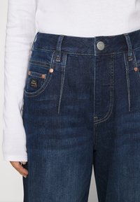 Mörkblå högmidjade jeans med koppardetaljer, synlig sömnad och en etikett på bakfickan. Texturerad denimtyg.