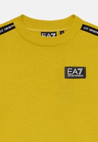Geltonas medvilninis marškinėliai su apvaliu iškirpimu, paveiksluojant juodą etiketę su "EA7 EMPORIO ARMANI" tekstu krūtinėje ir kontrastingomis pečių juostelėmis.