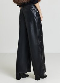 Schwarze Lederhose mit weitem Bein, versehen mit Druckknopfdetails entlang der Seiten und zwei Gesäßtaschen; kombiniert mit schwarzen Stiefeln mit Blockabsatz.