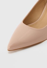 Chaussure plate à bout pointu en cuir verni nude brillant avec une semelle marron clair, vue en angle depuis le dessus.