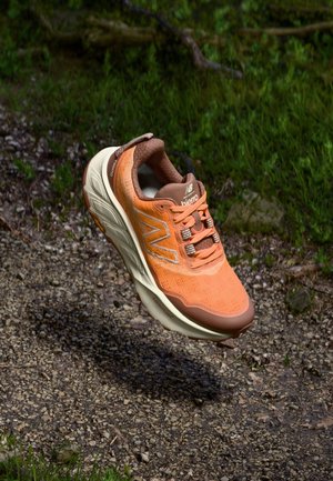 Chaussure de trail running orange avec une tige en mesh, des accents bruns et une semelle rembourrée. Présente des lacets et un motif texturé à la surface.