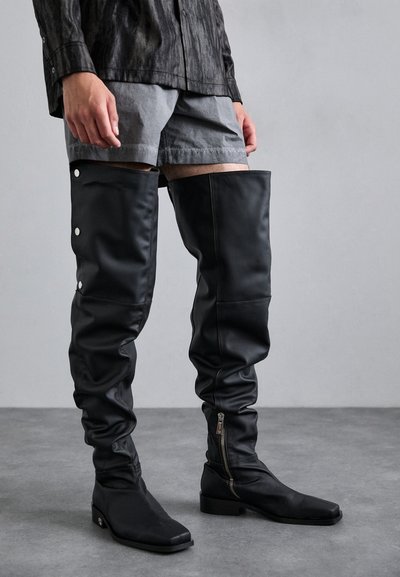 Schwarze Overknee-Stiefel aus strukturiertem Material mit glänzenden Akzenten. Mit einem seitlichen Reißverschluss und metallischen Druckknöpfen an der Außenseite.