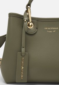 Zielona torebka ze skóry o fakturowanej powierzchni, z złotymi elementami, wyposażona w ozdobną metkę, górny uchwyt oraz regulowany pasek na ramię.