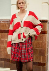 Ung kvinde med kort blond hår iført en rød og beige stribet cardigan og en rødternet nederdel, der står op ad en flisevæg.