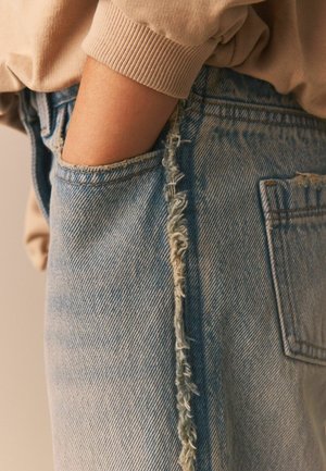 Bras dans un pull beige rangé dans la poche avant d'un jean en denim bleu clair avec un détail de couture verticale effilochée.