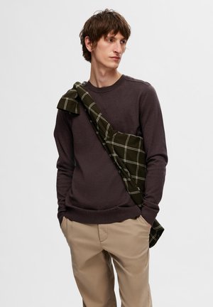 Maglione a maniche lunghe marrone scuro abbinato a pantaloni khaki, con una camicia a quadretti verde appoggiata sulla spalla.