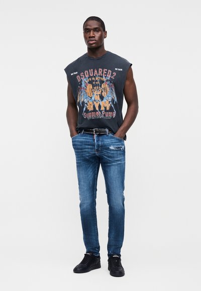 Homme debout, mains dans les poches, portant un débardeur noir avec un motif graphique de sport, un jean bleu déchiré, une ceinture noire et des baskets noires sur fond blanc.