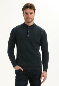 Man die een donkerblauwe gestructureerde langslavende gebuttonde trui draagt met één hand in zijn zak tegen een neutrale achtergrond.
