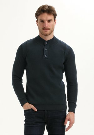Man die een donkerblauwe gestructureerde langslavende gebuttonde trui draagt met één hand in zijn zak tegen een neutrale achtergrond.