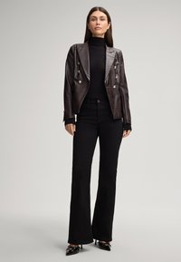 Bruine faux leren dubbelgeknoopte blazer met zilveren knopen, gedragen over een zwarte ribgebreide coltrui en zwarte flared jeans, geaccentueerd met puntige hakken.