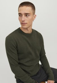 Pull en maille vert olive avec un col rond, des poignets côtelés et une texture lisse. La coupe semble slim et ajustée.