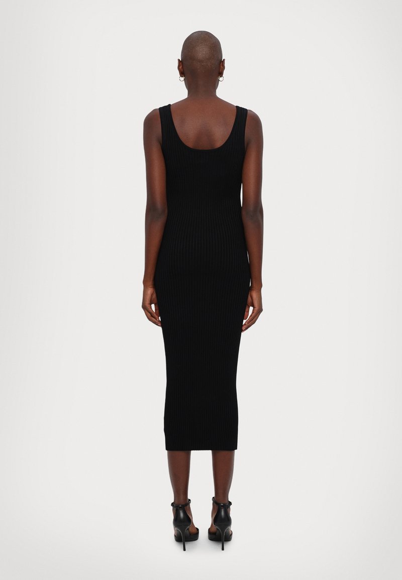 Femme avec la tête rasée portant une robe midi noire sans manches en maille et des sandales noires à talons hauts, se tenant de dos sur un fond uni.