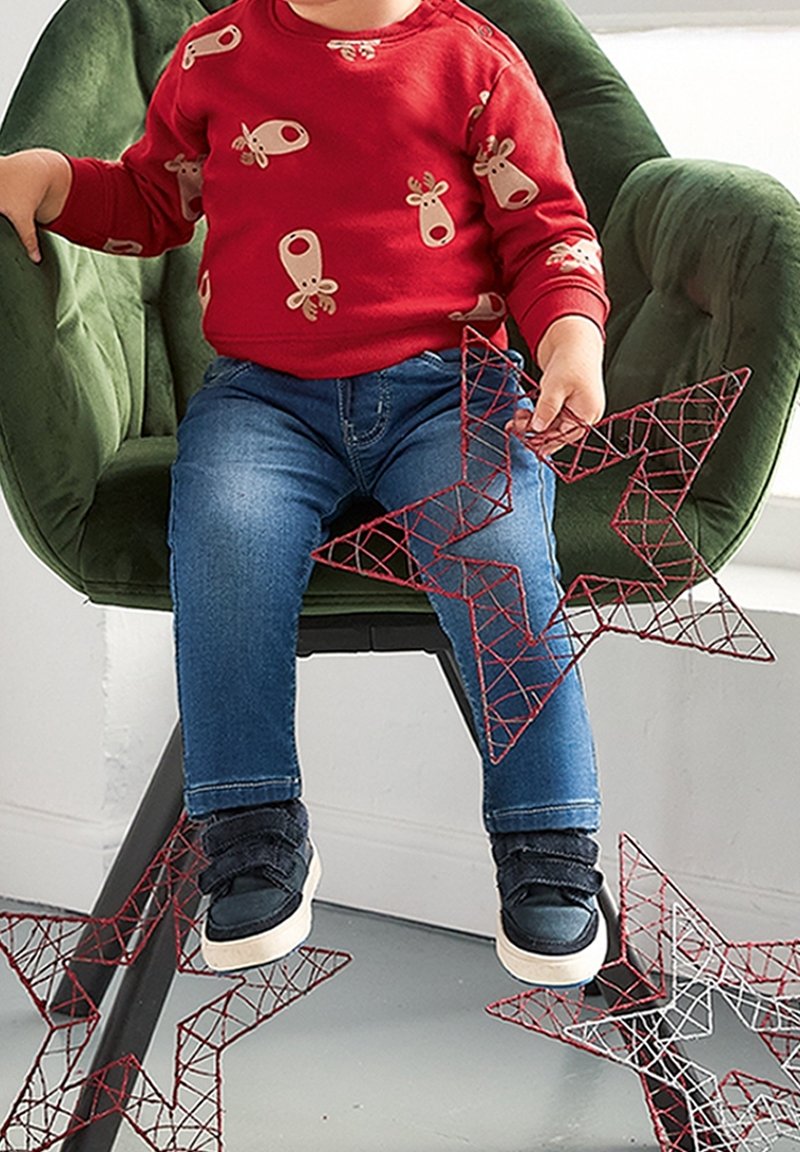 Tout-petit en pull renne rouge et jean assis sur une chaise verte tenant une étoile en fil rouge avec deux autres étoiles sur le sol gris.