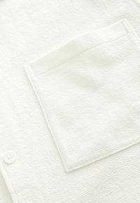 Chemise blanche texturée avec une fermeture à boutons, comportant une poche poitrine unique. Le matériau semble doux avec un motif subtil.
