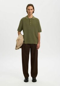 Jeune homme portant un polo à manches courtes vert olive, un pantalon marron foncé, des chaussures noires, tenant une veste beige sur un fond clair uni.