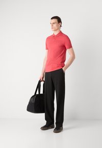 Koralna polo majica z dvema gumboma, črne široke hlače in temni športni copati. Drži črno torbo za trening z naramnicami. Minimalističen stil.