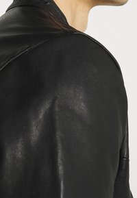 Černá kožená bunda s hladkou texturou, výrazným švem na rameni a klasickým límcem, zdůrazňující elegantní, moderní siluetu.