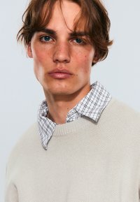 Suéter de punto beige superpuesto sobre una camisa de cuadros en blanco y negro, con un cuello clásico, textura suave y corte relajado.