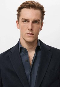 Bărbat într-un sacou închis la culoare peste o cămașă bleumarin cu dungi. Sacoul are un buzunar și revere, iar cămașa are un guler cu nasturi.