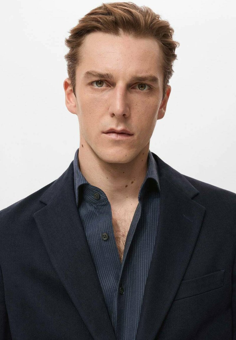 Mann in einem dunkelblauen Blazer über einem gestreiften Marinehemd. Der Blazer hat eine Tasche und Revers, während das Hemd einen Knopfkragen besitzt.