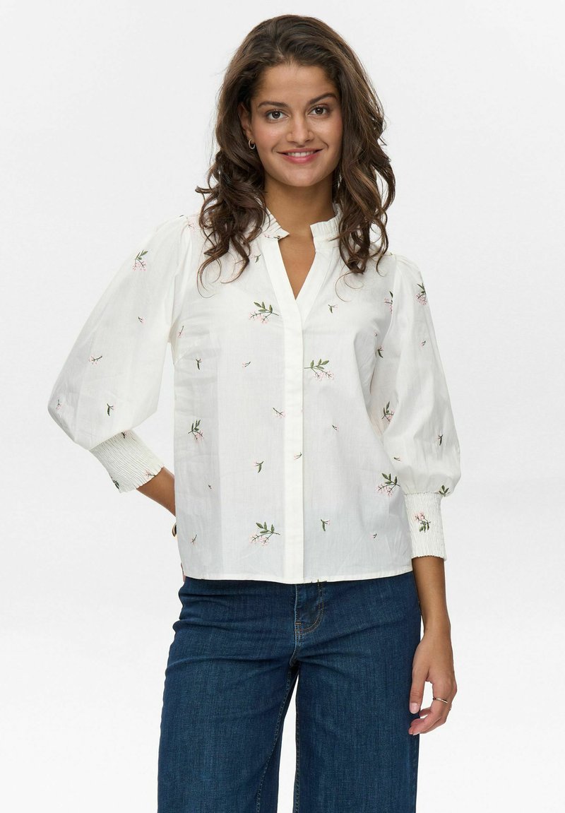 Blusa bianca con ricami floreali, maniche a sbuffo e scollatura arricciata. Abbinata a jeans blu scuro, che presentano una vestibilità comoda.