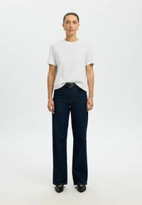 Femme debout, faisant face à l'avant, portant un t-shirt blanc à manches courtes, un jean bleu marine à jambes larges avec ceinture, et des chaussures noires à bout pointu.