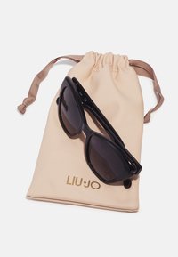 Lunettes de soleil noires au design en œil de chat pointu reposant sur un sac à cordon beige avec un logo doré "LIU·JO".