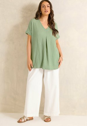 Vrouw draagt een lichtgroene getextureerde V-hals tuniek, witte wijde broekspijpen en beige geweven sandalen, staand tegen een beige achtergrond.