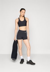 Črn športni crop top in kratke hlače iz raztegljivega materiala. Model drži črno brisačo, nosi črne športne copate in bele nogavice do gležnjev.