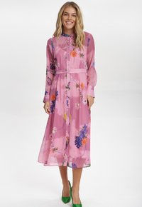 Rosa blommig klänning med genomskinligt tyg, långa ärmar, knappslå framtill, midja med bälte och flerfärgade blommönster. Avslutad med gröna klackar.