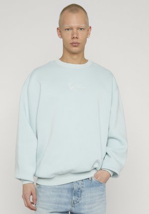 Giovane con capelli corti che indossa una felpa oversize azzurro chiaro e jeans azzurri chiari, in piedi con una mano in tasca.