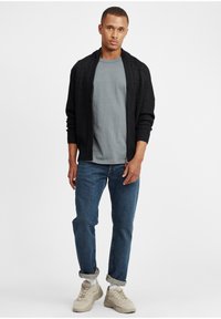 Cardigan nero a maglia sopra una maglietta grigia, abbinato a jeans blu e sneaker beige. Il cardigan presenta un motivo testurizzato e apertura frontale.