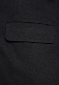 Černá tkanina se vodorovnou kapsou s klopou. Textura je hladká a mírně elastická, typická pro pletený materiál.
