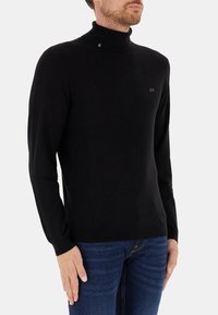 Maglione nero a collo alto realizzato in tessuto morbido, con un design aderente, polsini a costine e un piccolo logo sul petto. Abbinato a jeans blu.