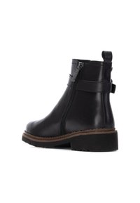 Botas de piel negra hasta el tobillo con cremallera lateral, suela de goma marrón plana y tacón apilado. Presenta un bucle para tirar en la parte posterior.