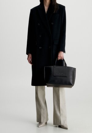 Sort dobbeltradet frakke, lysegrå vidde bukser, beige spidse hæle og en sort læder shopper med præget Calvin Klein-logo.