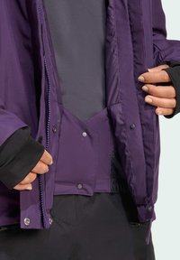 Purppura eristetty takki, jossa on vetoketjullinen etuosa ja nepparikiinnitys, harmaa sisävuori ja mustat hihansuut. Kestävä kankaan tekstuuri.