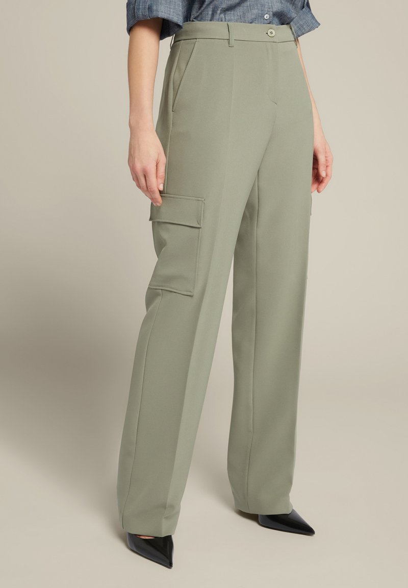 Olijfgroene wijde broek van soepel stof, voorzien van zijzakken en een hoge taille, gecombineerd met zwarte puntige hakken.