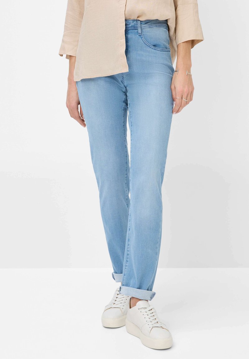 Jeans de mezclilla azul claro con corte recto, cintura media y puños enrollados, combinados con zapatillas blancas y una blusa beige.