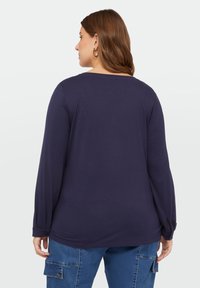 Marineblauw langemouwtop met een ronde halslijn, iets losse pasvorm, knoopdetail bij de mouwmanchetten, gemaakt van zachte stof. Gedragen met denim broek.
