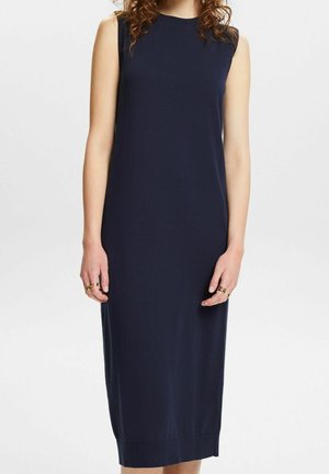 Kobieta ubrana w granatową, bezrękawnikową sukienkę do kolan z okrągłym dekoltem, stojąca na tle jednolitego, jasnego tła.