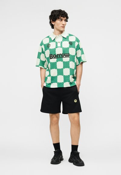 Jeune homme portant une chemise à manches courtes à carreaux verts et blancs, un short noir avec un patch smiley jaune, des chaussettes noires et des baskets noires, debout immobile.