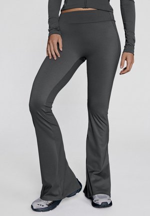 Personne portant un pantalon de sport évasé gris foncé et des chaussures de course grises et blanches, debout avec une jambe légèrement pliée.