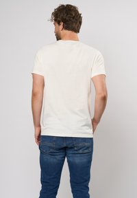 Kremfarget bomulls t-skjorte med korte ermer, rund hals, og et rent design, båret med blå denim jeans som viser stingene på baklommen.