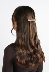 Fermaglio per capelli dorato arricciato che tiene assieme capelli castani ondulati, con una texture liscia e una finitura lucida, acconciati in uno stile mezzi raccolti.