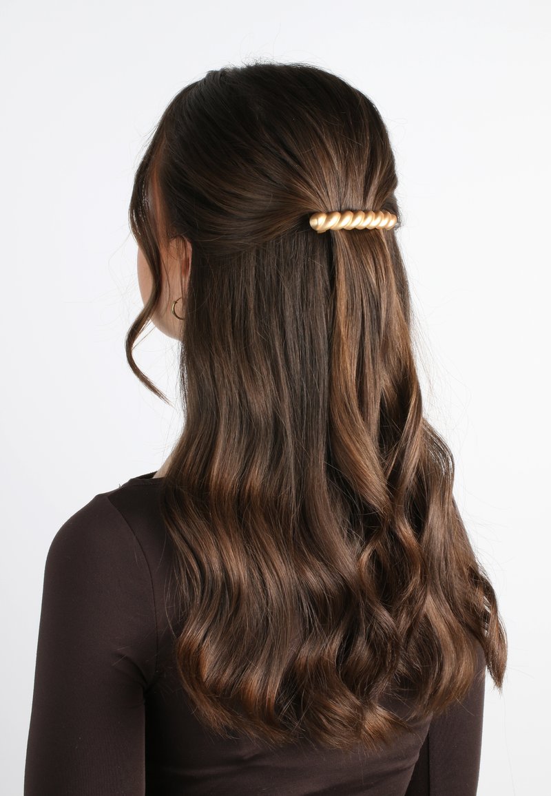 Fermaglio per capelli dorato arricciato che tiene assieme capelli castani ondulati, con una texture liscia e una finitura lucida, acconciati in uno stile mezzi raccolti.