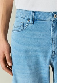 Main reposant à côté de la zone de la taille d’un jean en denim bleu clair, montrant la poche avant, la boucle de ceinture et les détails de la fermeture bouton.